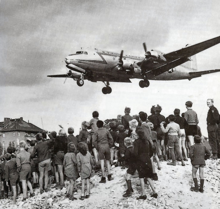 Avión Douglas C-54 Skymaster aterrizando en el aeropuerto de Tempelhof durante el puente aéreo de Berlín (1948-1949), con berlineses observando desde un montón de escombros.